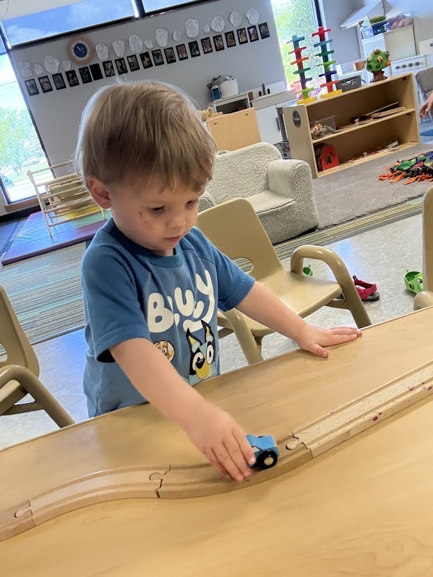Young child pushes a toy vehicle along a wooden train track during classroom play.