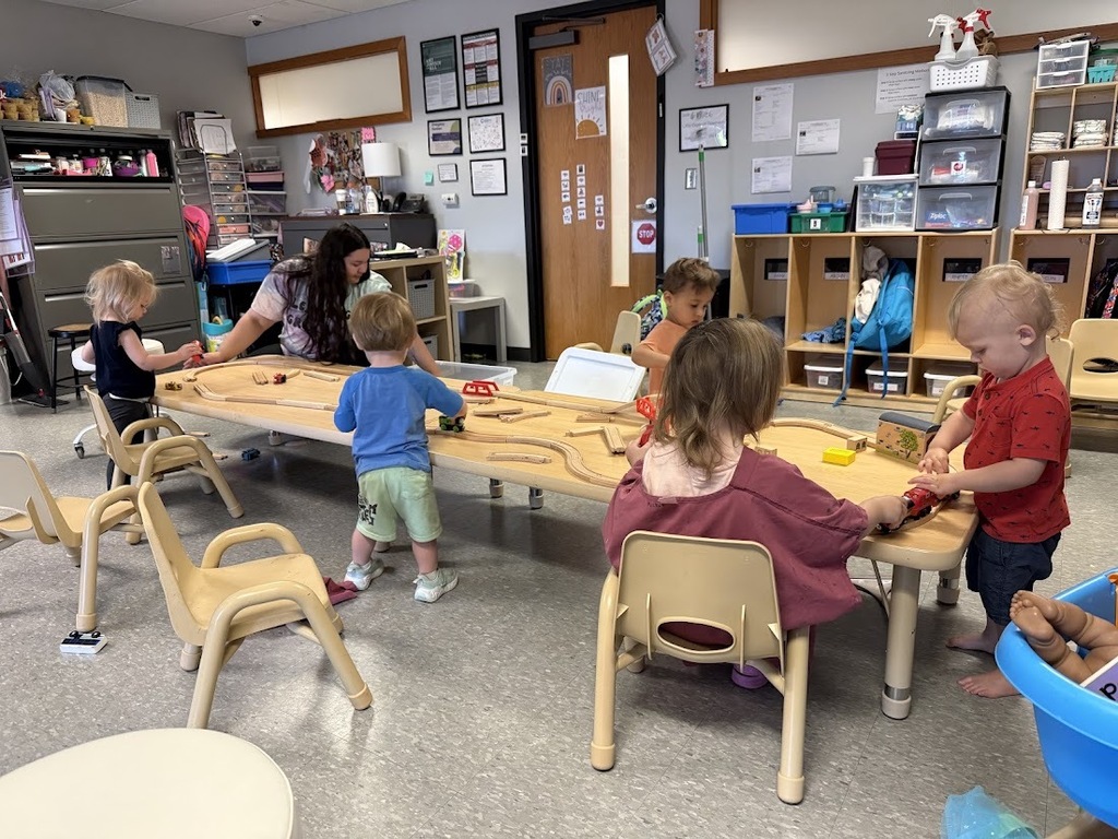 Several young children gather around train tables, building and playing with wooden train tracks together.