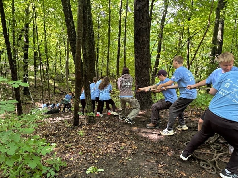 Group of youth working together to pull a thick rope during a game of tug-of-war in a dense, wooded forest.