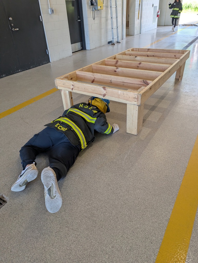 A person in full firefighter turnout gear and a helmet crawling under a low wooden structure inside a fire station bay.