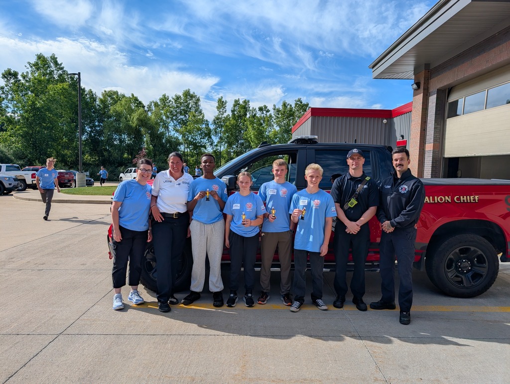 A group of youth holding small trophies stand alongside fire department staff for a group photo in front of a red Battalion Chief truck.