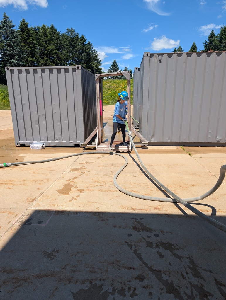 A youth wearing a blue shirt and hard hat maneuvers a large hose through a wooden doorway between two grey shipping containers during an outdoor activity.