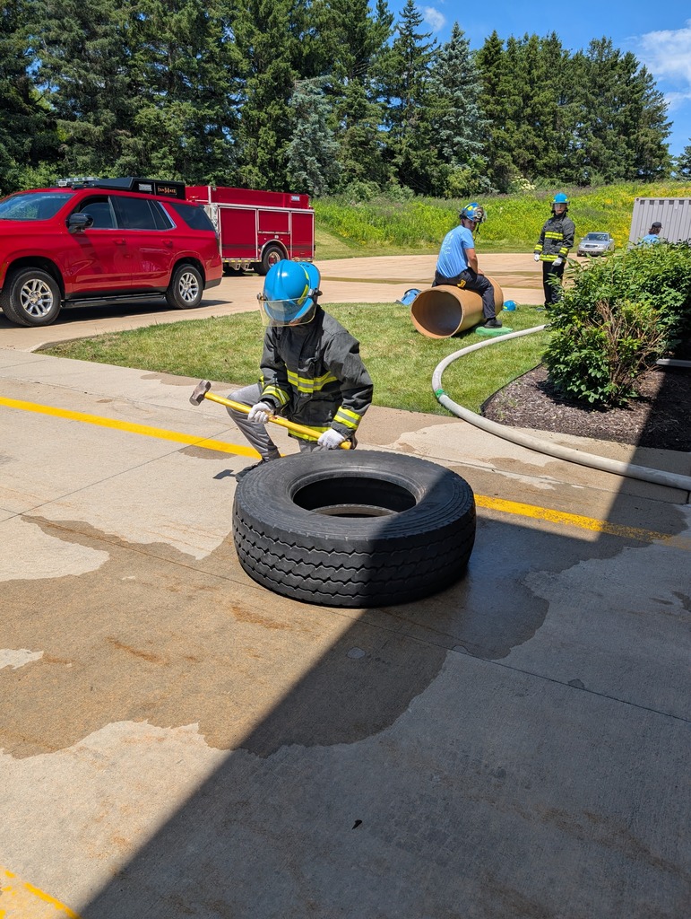 A person in a firefighter jacket and blue helmet holds a sledgehammer next to a large tire during an outdoor training course, with fire vehicles in the background.