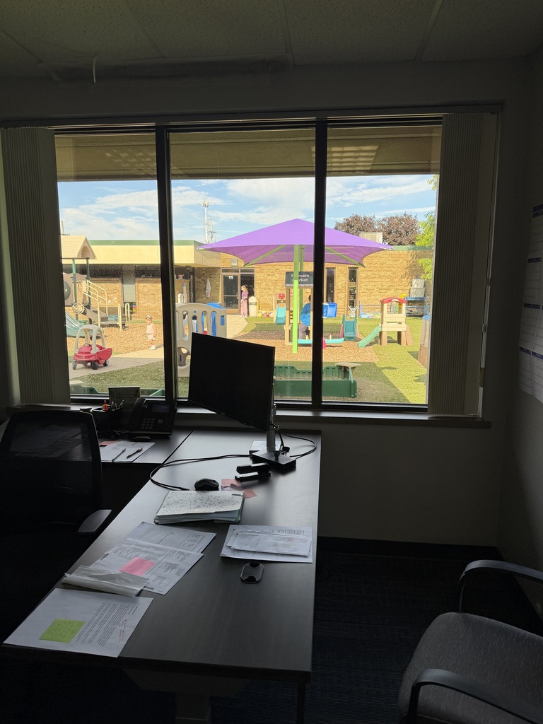 An office desk with a computer monitor, papers, a computer mouse, a stapler, and an office chair is in front of a window. Outside the window is a playground with a purple umbrella over a playset