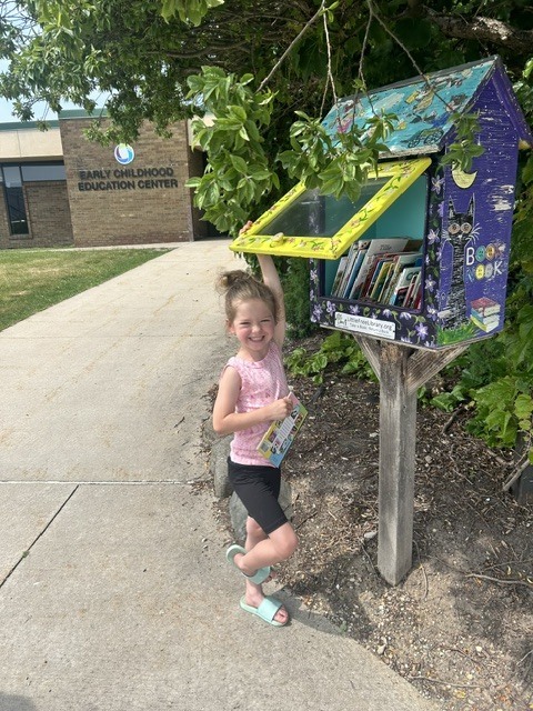 A young girl with her hair in a bun stands on a sidewalk, smiling brightly at the camera while holding up the yellow glass door of a colorful, hand-painted "Little Free Library" book nook. She is wearing a pink sleeveless shirt and black shorts, holding a children's book in her other hand. In the background, the brick building reads "Early Childhood Education Center."