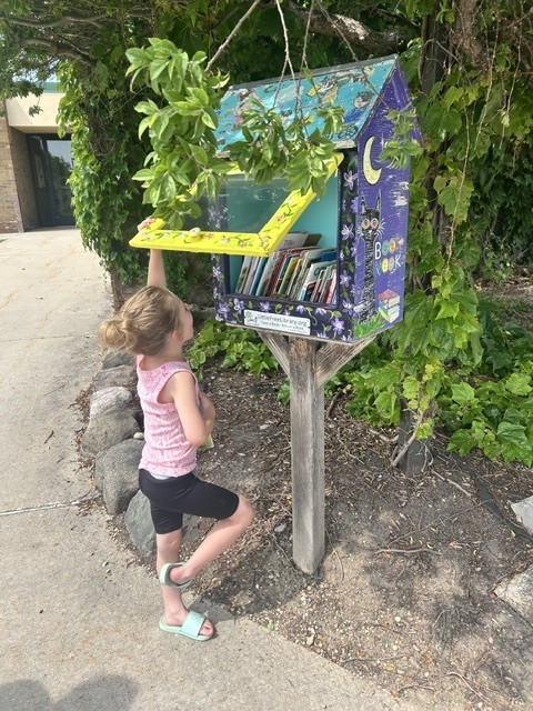 A young girl with her hair in a bun stands on a sidewalk, looking inside a colorful, hand-painted "Little Free Library" box mounted on a wooden post. She is holding the yellow glass door open with one hand while checking out the books inside.
