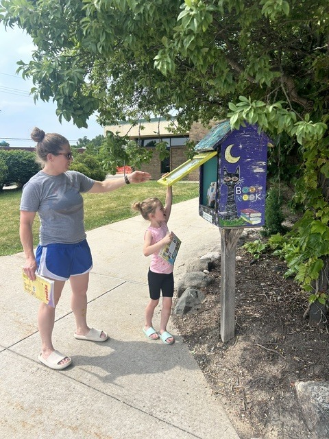 A woman and a young girl stand on a sidewalk next to a colorful, hand-painted "Little Free Library" box. The woman, wearing a grey t-shirt and blue shorts, holds a book and assists the girl, who is reaching up to hold open the yellow glass door of the book sharing box.