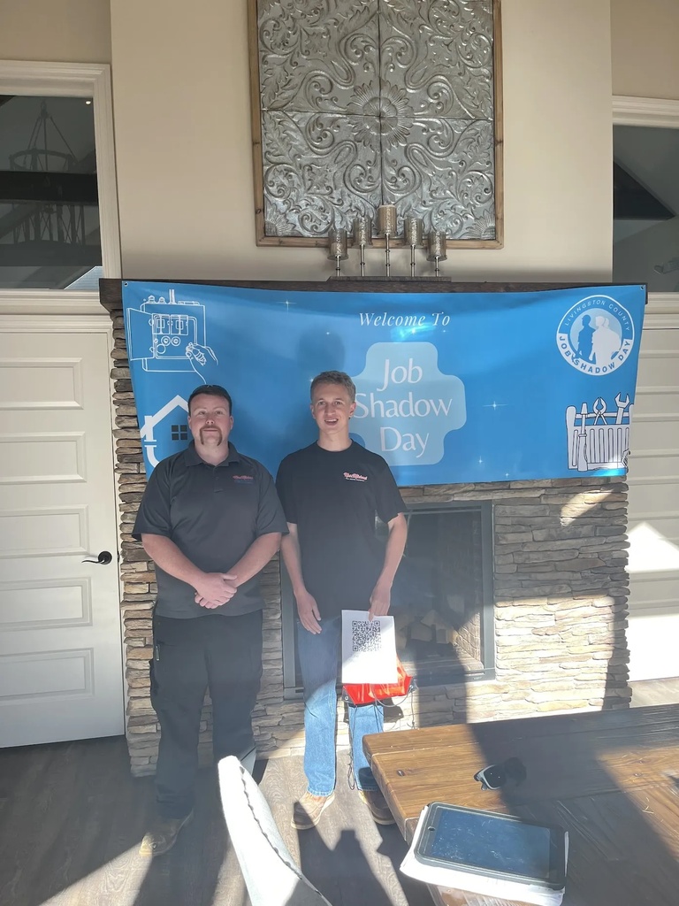 Two men standing together in front of a stone fireplace with a blue banner that reads "Welcome to Job Shadow Day."