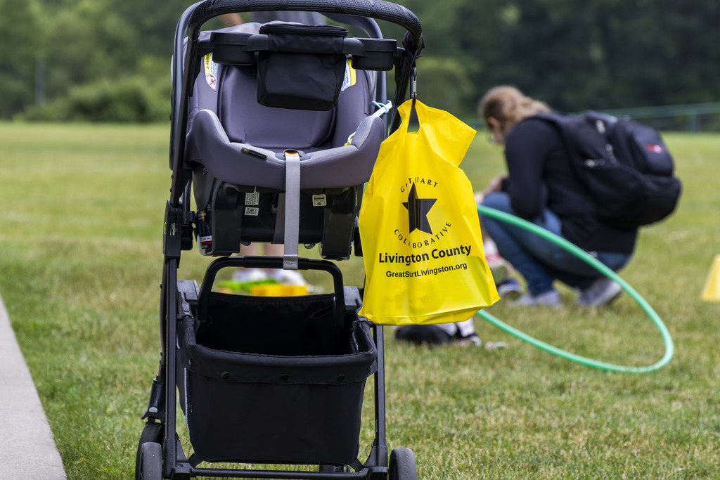 An empty stroller sitting on the grassy park ground with a bright yellow bag hanging from the handle. The bag reads "Great Start Collaborative Livingston County. GreatStartLivingston.org." A woman can be seen in the background crouching down.