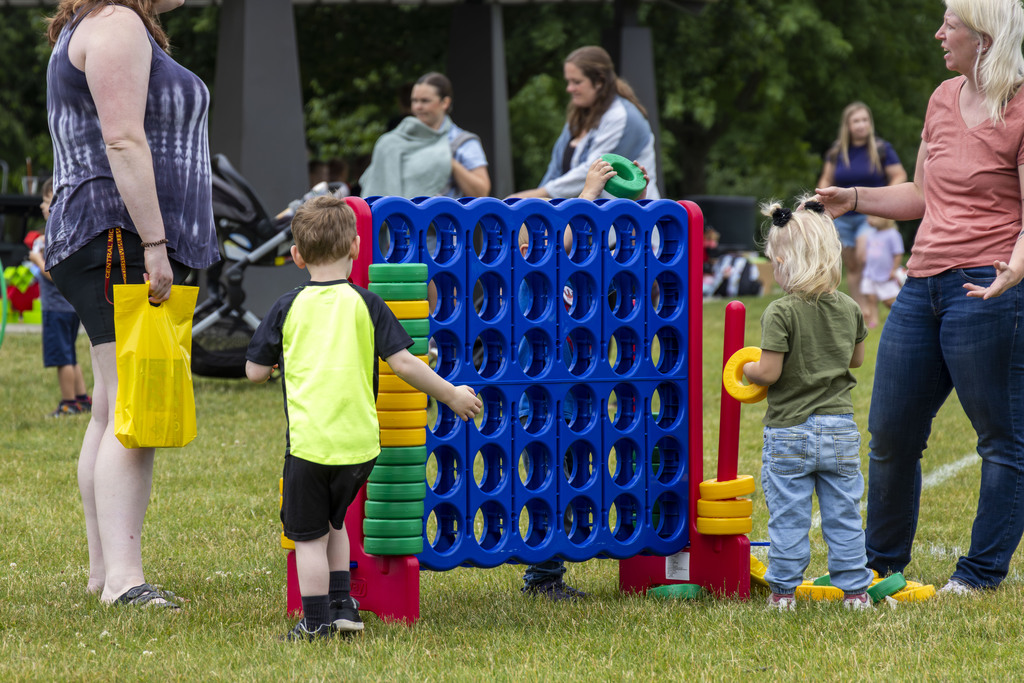 Two young kids are playing a giant game of connect four with yellow and green coins. Two parents are standing on either side chatting with one another.