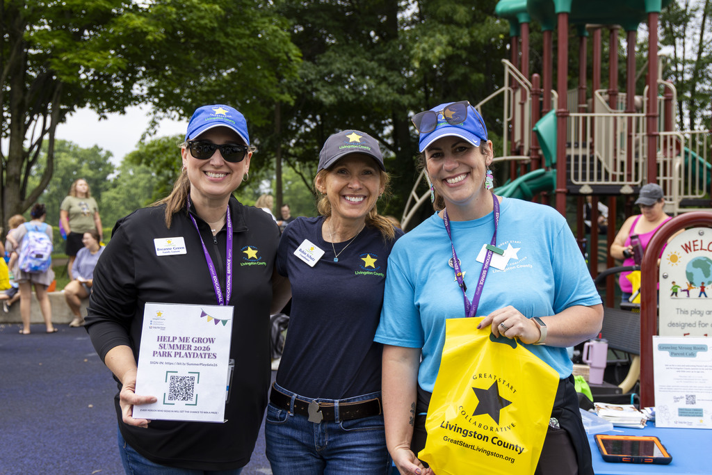 Three woman standing together. One woman is holding a bag that says "Great Start Collaborative Livingston County." Another is holding a QR code to register for "Summer 2026 Park Playdates."