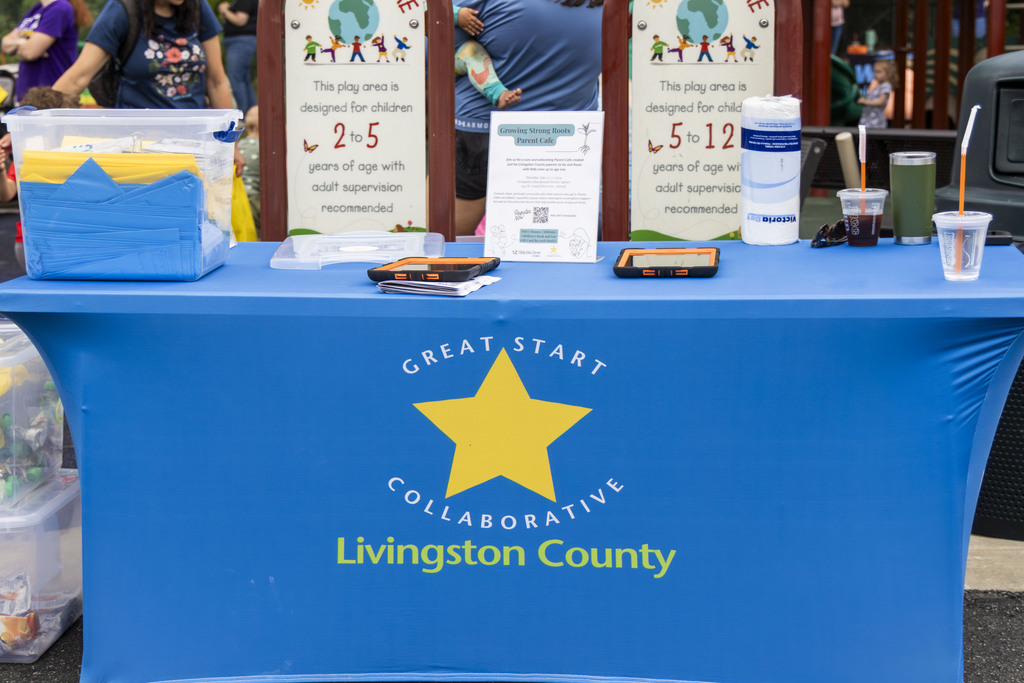 An almost empty table with a table covering that reads "Great Start Collaborative Livingston County." A few half-consumed beverages sit atop the table as well as a bin of stacked yellow and blue bags.