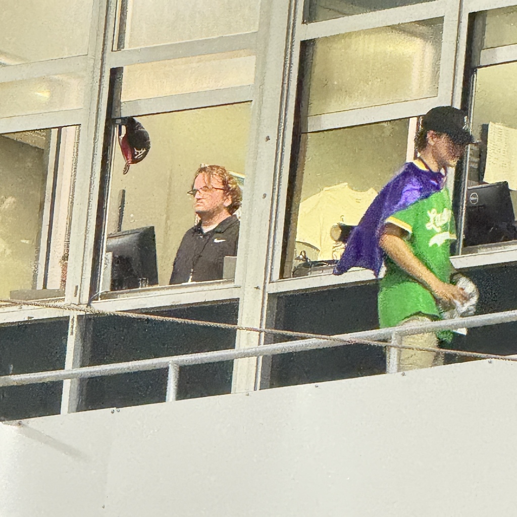 A view from outside a stadium press box or booth windows. Inside, a man with glasses and wavy hair looks out toward the field, while a person wearing a green jersey and a purple cape stands nearby on an outdoor walkway.