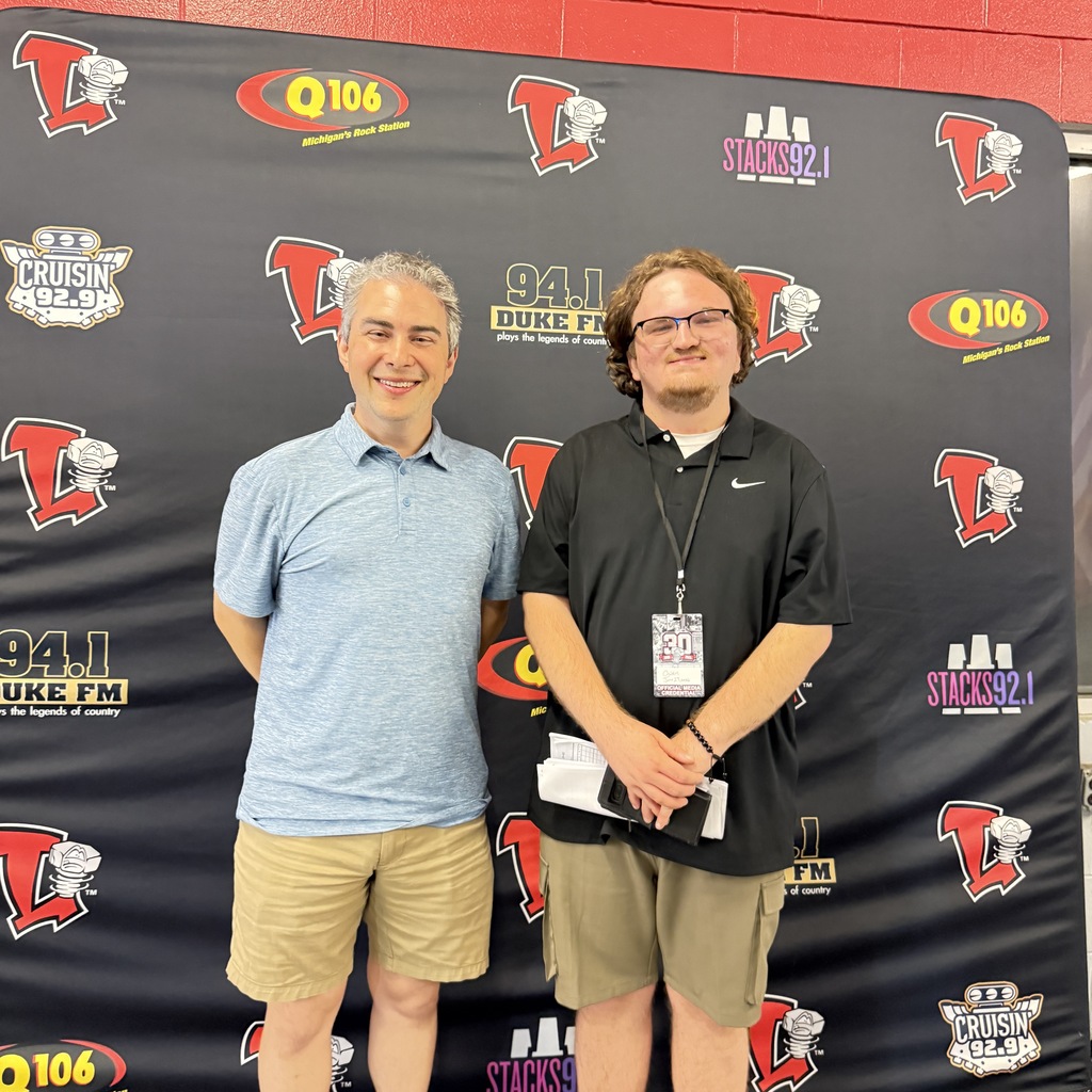 Two men stand side-by-side smiling in front of a black backdrop covered in local radio station logos and Lansing Lugnuts team logos. The man on the right wears a black polo shirt, khaki shorts, and a media credential lanyard.
