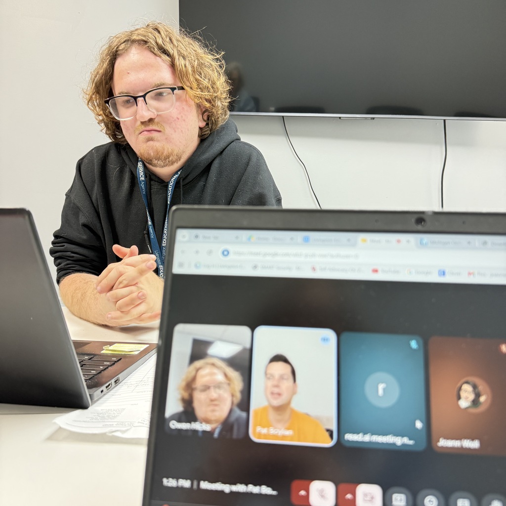 A close-up view focused on a Google Meet video call on a laptop screen, showing participants including a man named Pat Boylan. In the background, the same young man with wavy hair and glasses sits with his hands clasped, watching the meeting.