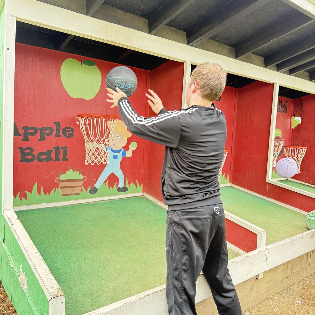 A close-up shot from behind a young man in a black tracksuit as he shoots a black basketball toward a low hoop in the "Apple Ball" game booth.