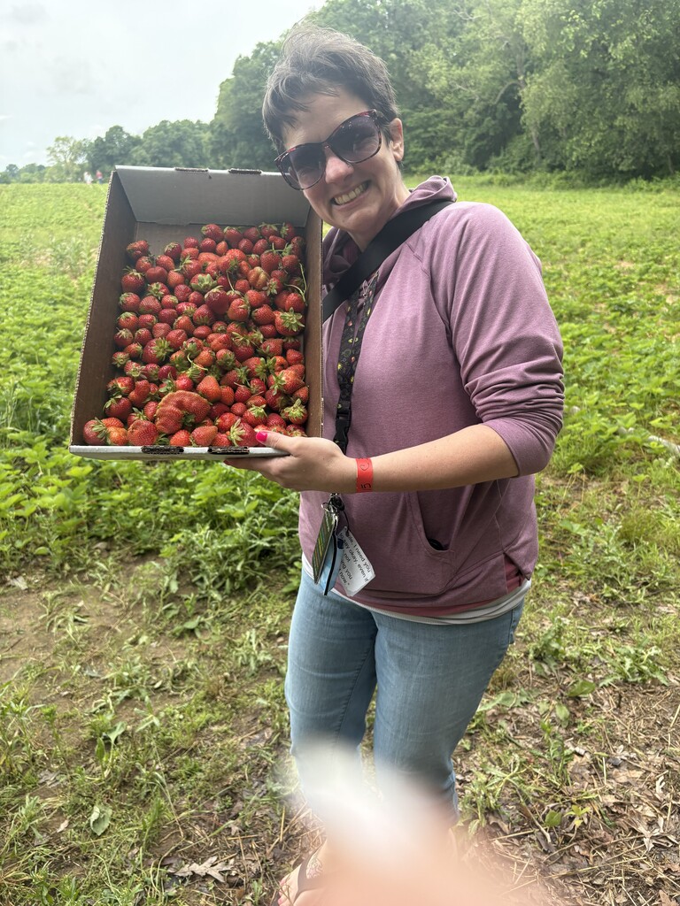 A smiling woman with short hair and sunglasses stands in a field, holding up a large cardboard box completely filled to the brim with freshly picked red strawberries.
