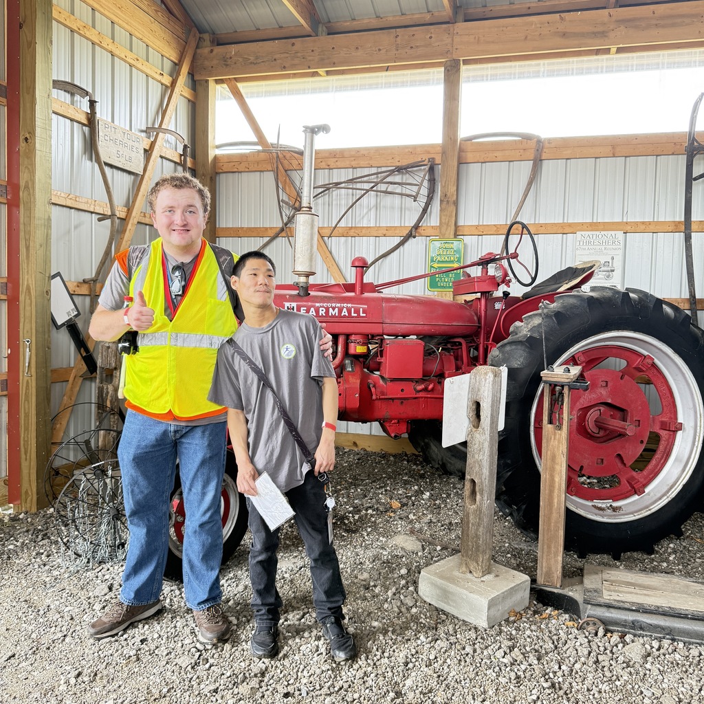 Two men pose inside a rustic barn in front of a vintage red McCormick Farmall tractor. The man on the left gives a thumbs-up while wearing a bright yellow safety vest.