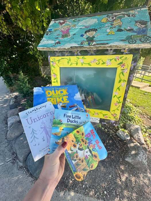 A hand holding four books in front of a little library.