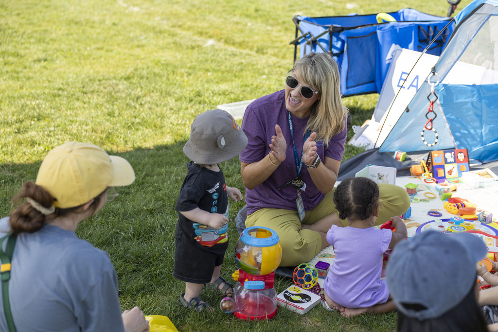 A smiling woman in a purple shirt claps while sitting on the grass with toddlers and baby toys during an outdoor community event.