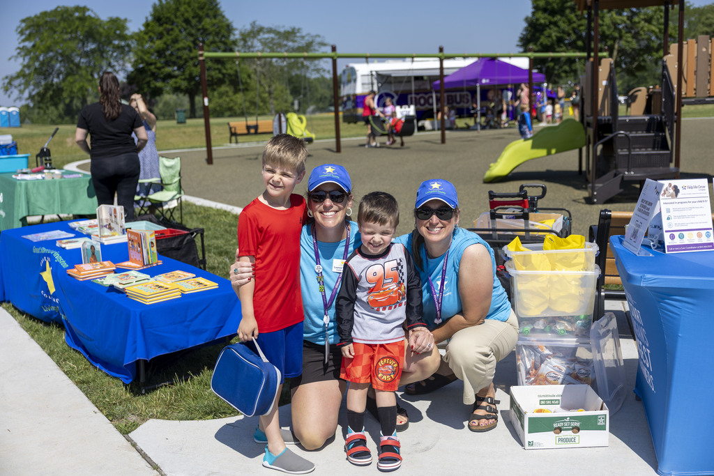 Two women in matching teal shirts and blue caps pose with two smiling young boys at a park playground near a book table.