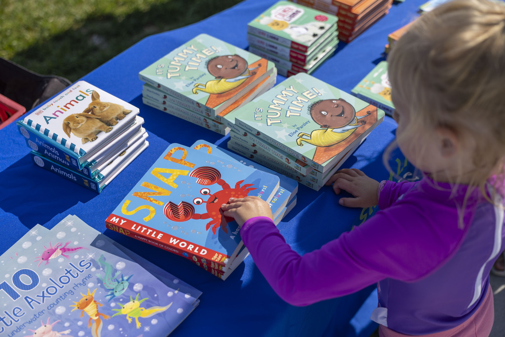 A young girl with blonde hair reaches for colorful children's board books stacked on a table covered in a blue tablecloth.