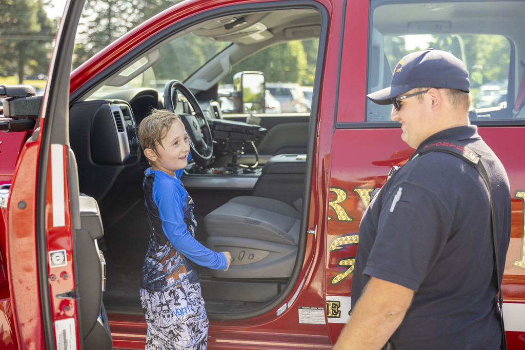 A young boy in a blue swim shirt smiles as he stands in the open door of a red fire utility truck next to a friendly firefighter in uniform.