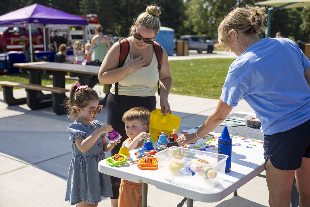 Two young children and a woman explore colorful toy musical instruments on a white folding table outdoors, assisted by a volunteer in a blue t-shirt.