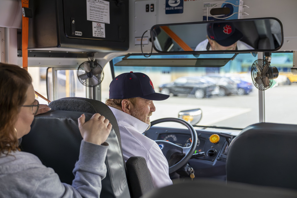 A school bus driver wearing a baseball cap smiles and chats with a woman sitting behind him.