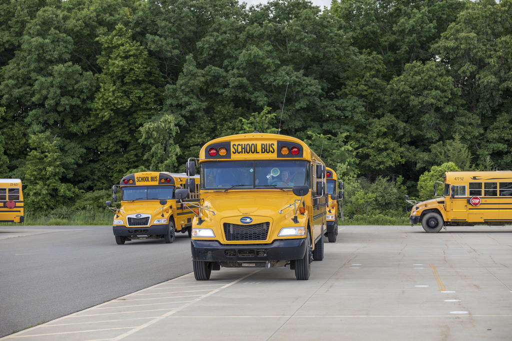 Several yellow school buses are parked on a paved lot in front of a dense line of green trees, with one driver waving a green smiley face sign.