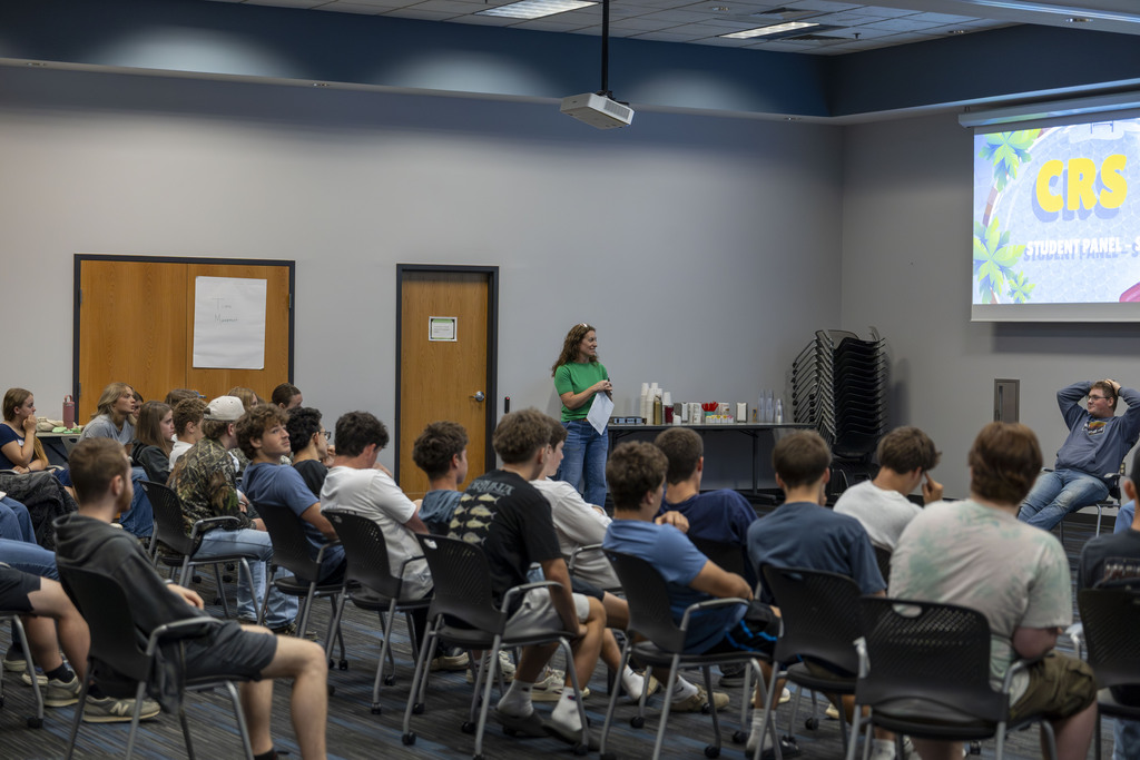 A group of students sitting in rows of chairs facing a presentation screen while a woman in a green shirt speaks to them.