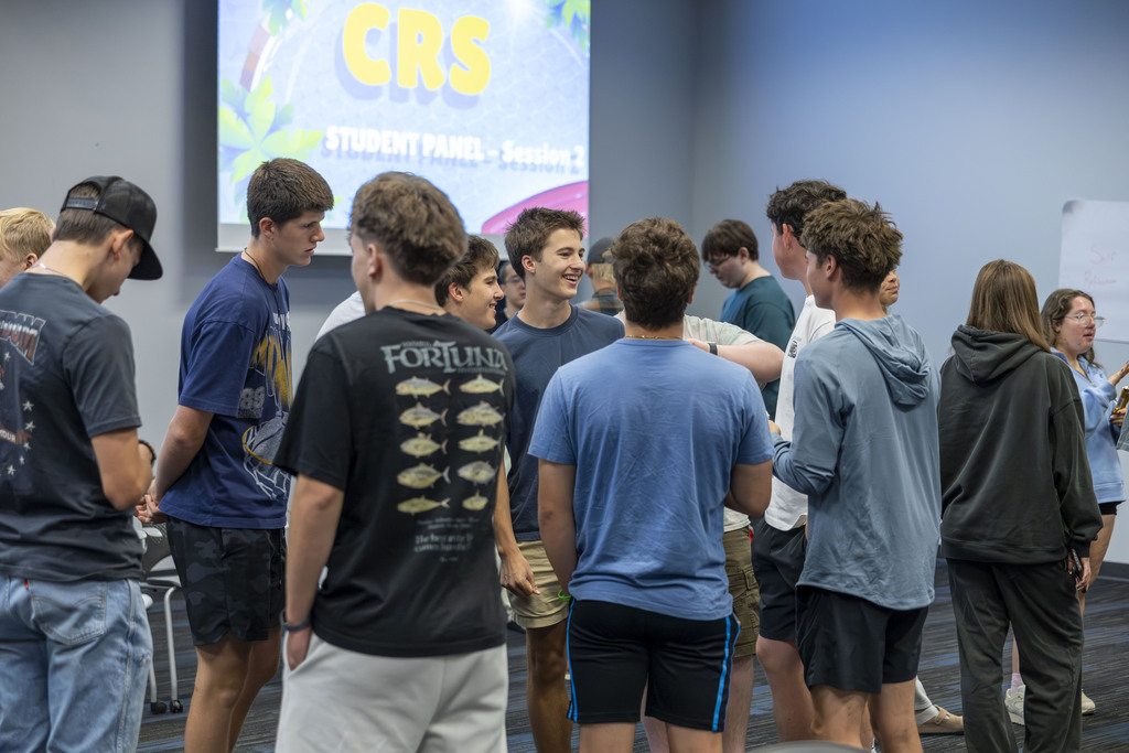 A group of young men standing in a circle and talking under a presentation screen.