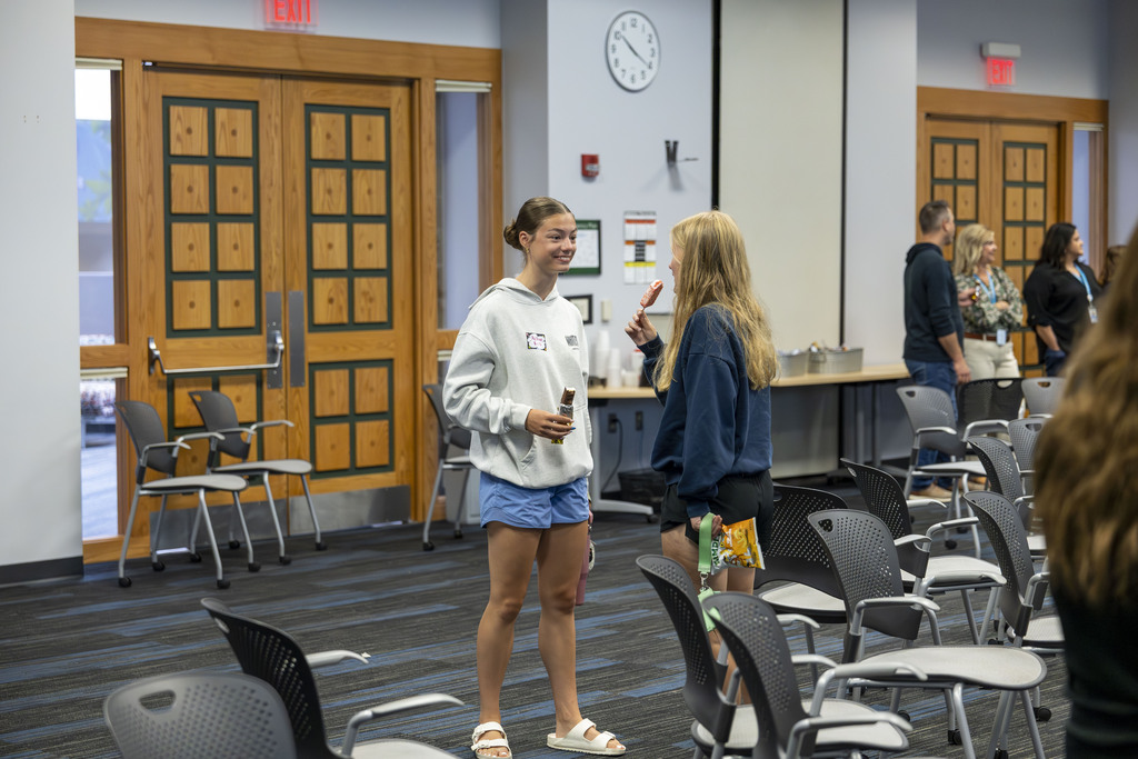 Two teenage girls stand in a large classroom, talking and eating ice cream treats.