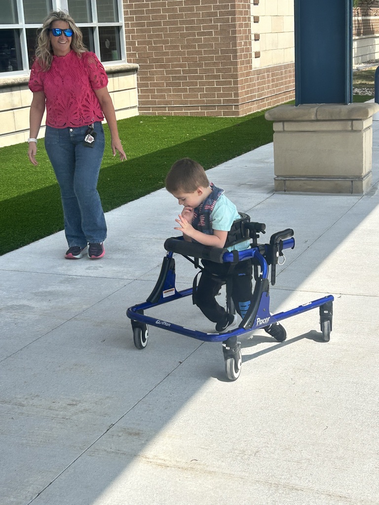 A young boy uses a blue adaptive walker on a sidewalk while an adult supervisor watches nearby.