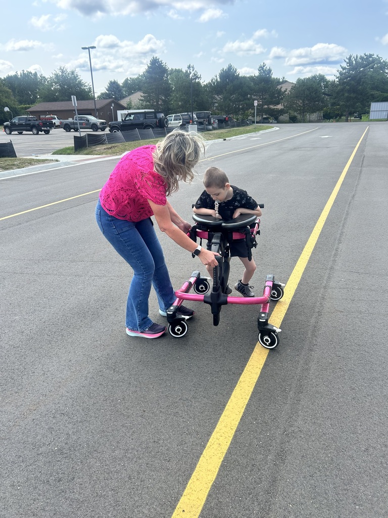 A woman assists a young boy as he uses a pink adaptive mobility walker on an asphalt road.
