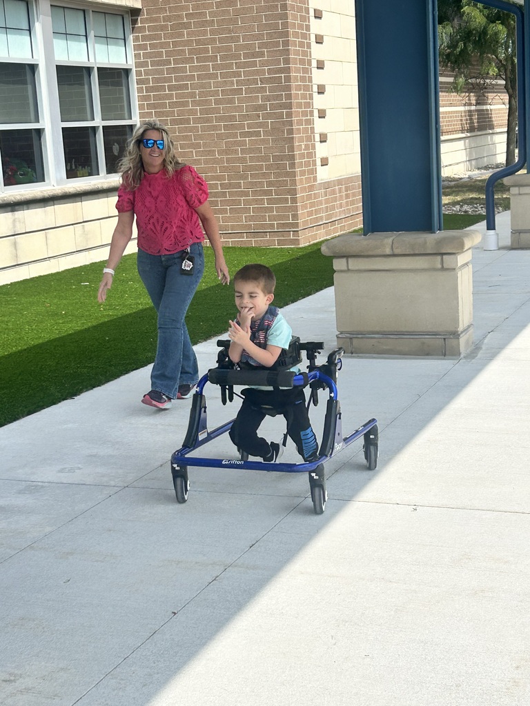 A young boy smiles as he moves along a sidewalk in a blue gait trainer, followed closely by a woman in a bright pink shirt.
