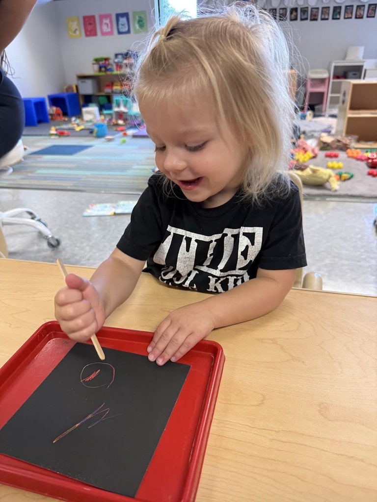 A smiling young girl with blonde hair uses a wooden stylus to draw on rainbow scratch paper in a classroom.