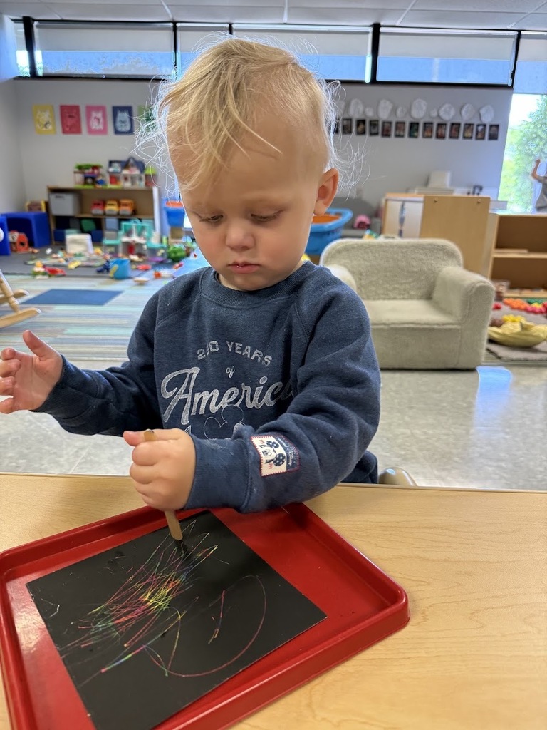 A blonde toddler in a navy sweatshirt focuses on making colorful scratch-art lines at a table.