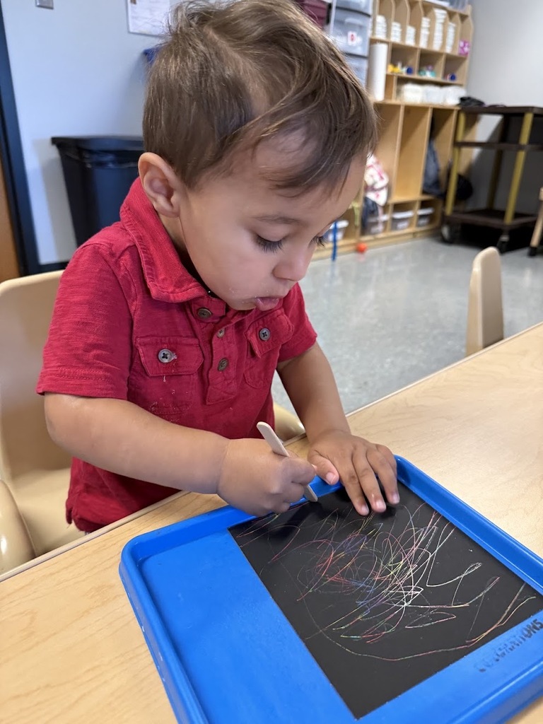 A focused toddler in a red shirt scratches colorful scribbles onto black paper using a wooden stick.