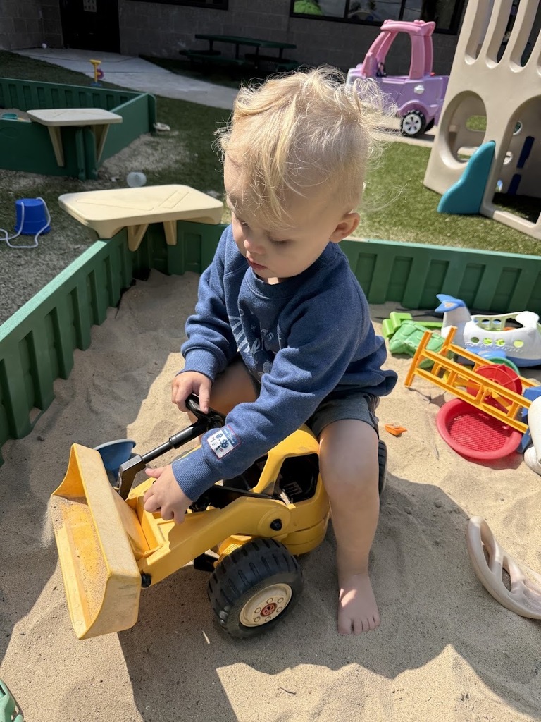 A toddler straddles and plays with a large yellow toy bulldozer inside a sandbox.
