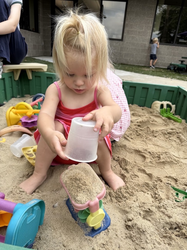A young girl in a pink and red sundress uses a plastic cup to build in the sand.