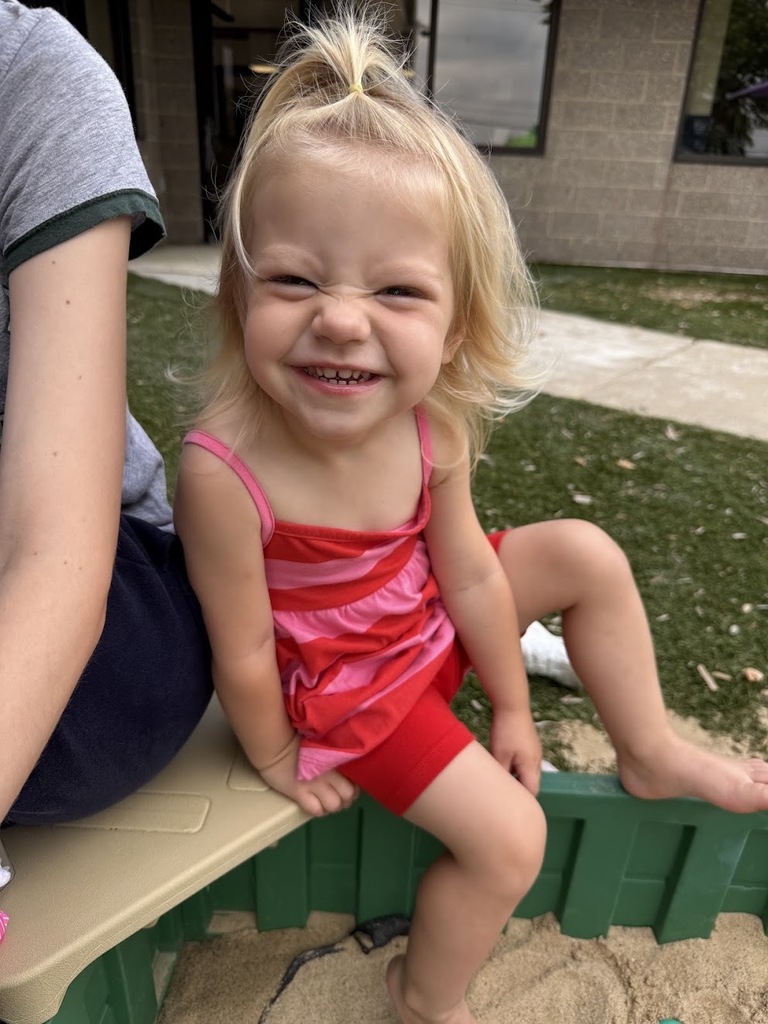 A smiling toddler girl with blonde hair tied in a topknot scrunches her nose joyfully in an outdoor sandbox area.