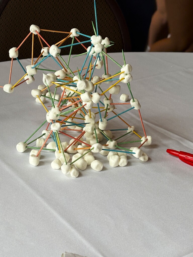 A close-up view of a complex structure built out of a mini marshmallows and colorful toothpicks resting on a white tablecloth.
