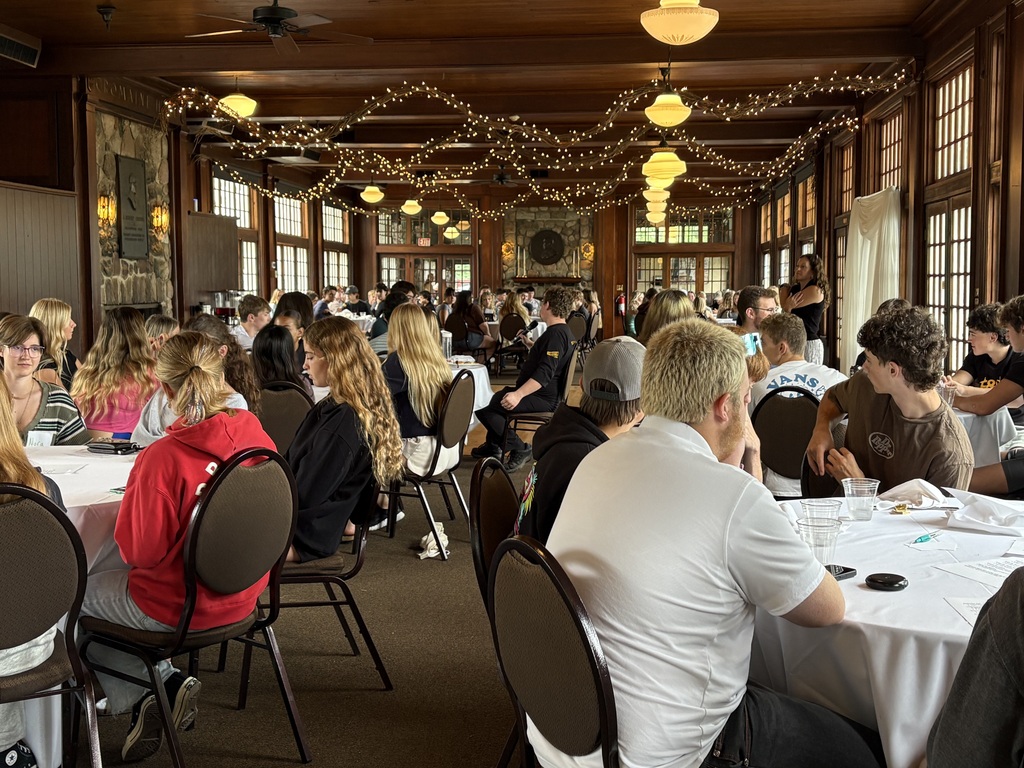A large group of people seating around round dining tables in a wood-paneled hall decorated with strong lights.