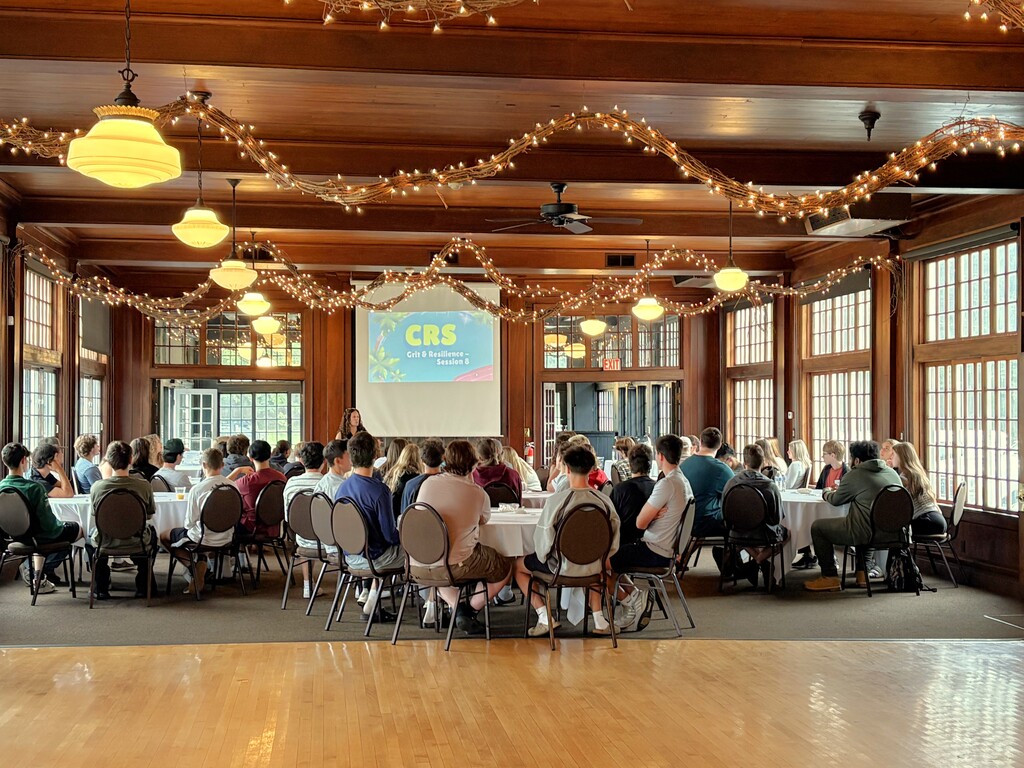 An audience seating at round tables faces a presentation screen reading "CRS Grit & Resilience" inside a wooden hall decorated with string lights.