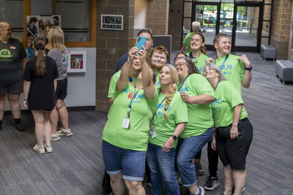 A group of volunteers in matching green t-shirts huddle together to take a group selfie in a hallway.