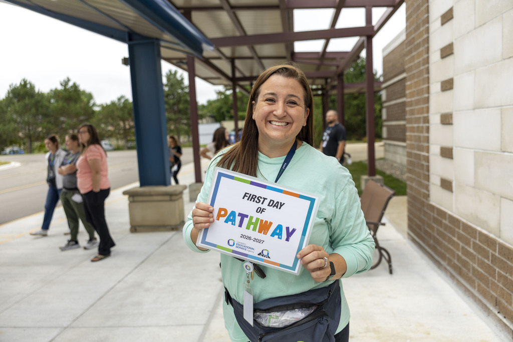A woman in a mint green shirt smiles outdoors while holding a "First Day of Pathway" sign under a covered walkway.