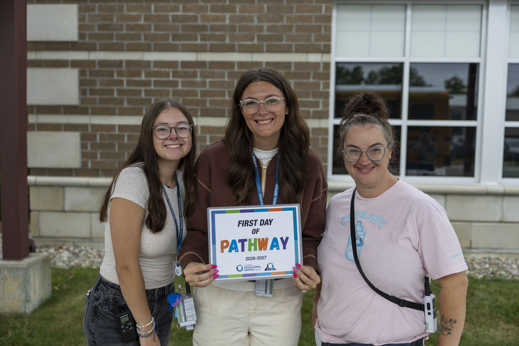 Three women pose together holding a "First Day of Pathway" sign outside a school building.