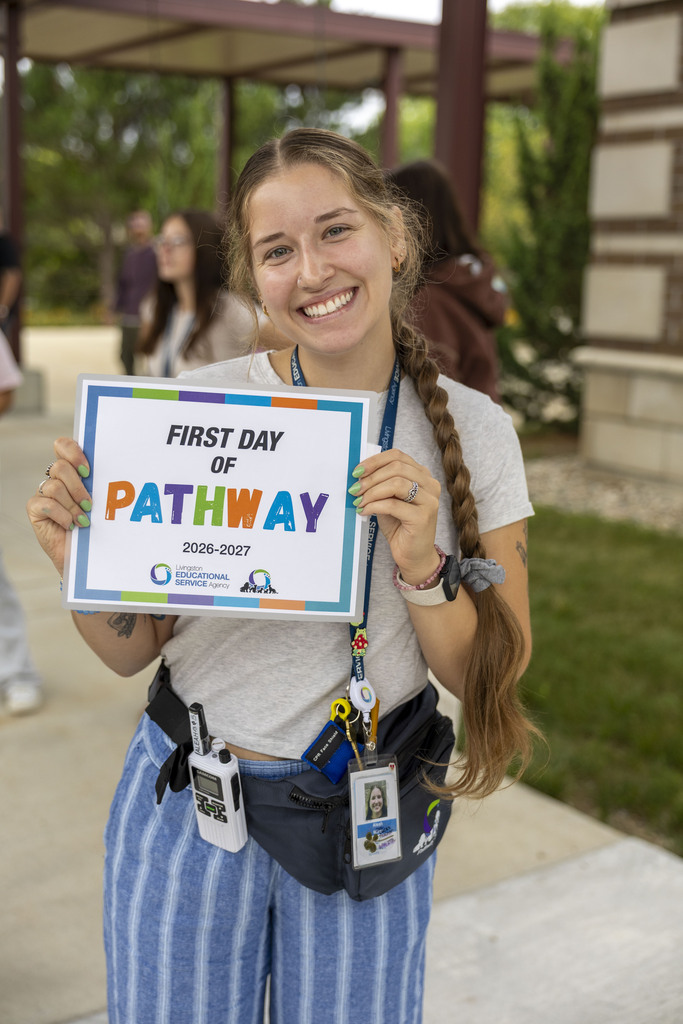 A young woman with a long side braid smiles as she holds a "First Day of Pathway 2026–2027" sign outdoors.