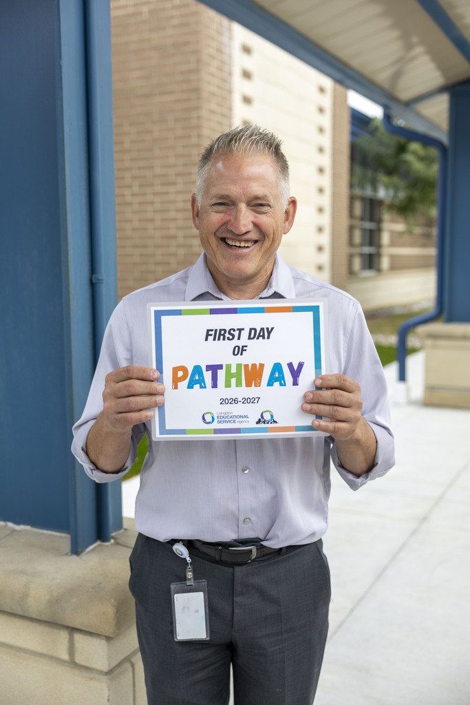 A smiling man in a button-down shirt and grey slacks holds a "First Day of Pathway" sign outdoors.